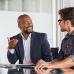 Two happy business colleagues at meeting in modern office interior. Successful african boss in a conversation with young employee in boardroom. Marketing team of two businessmen discussing strategy in meeting room.